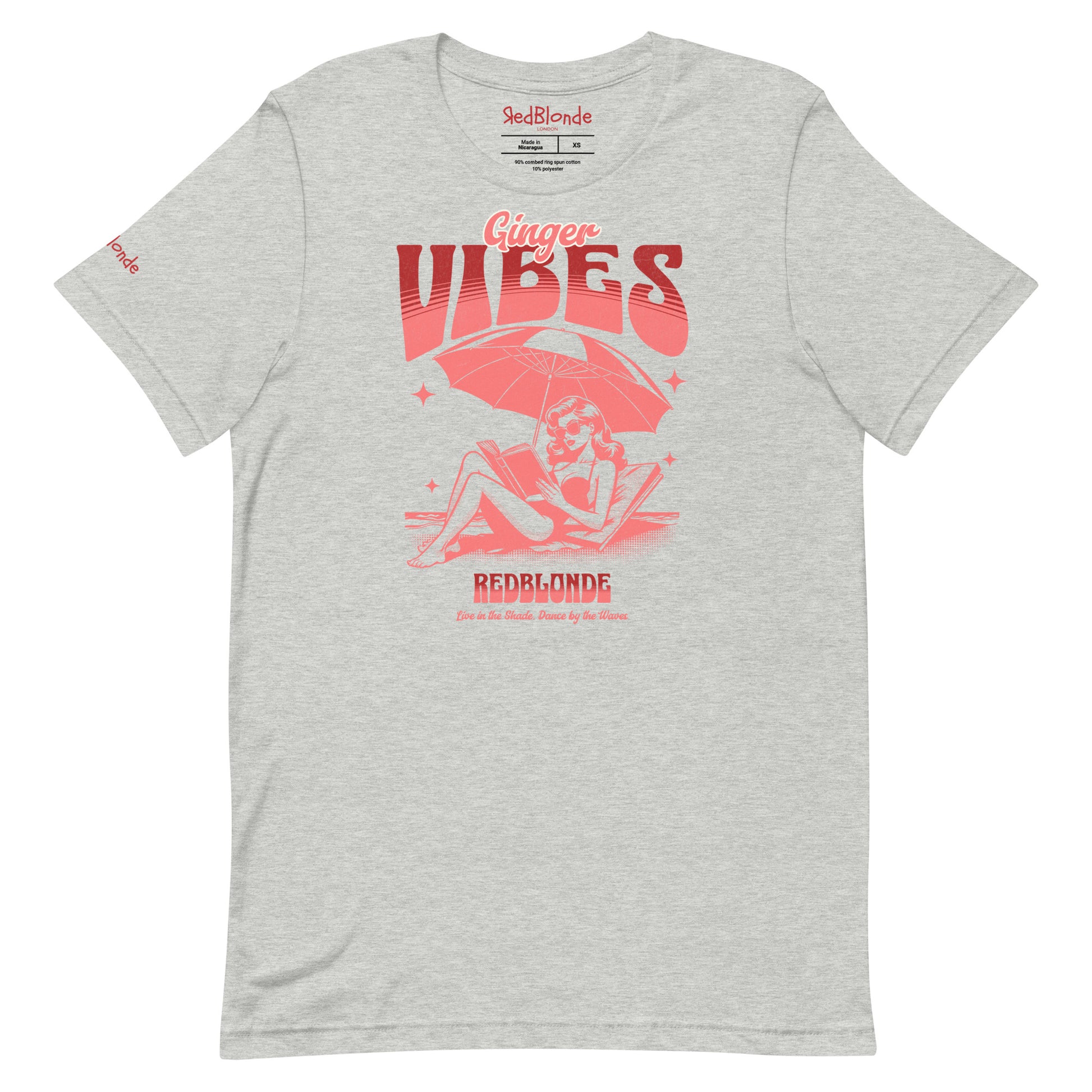 grey unisex t-shirt featuring a retro-style red illustration of a redhead woman lounging under a beach umbrella reading a book, with bold text reading "Ginger Vibes" above and "RedBlonde" below, alongside the tagline "Live in the shade. Dance by the waves." Branded RedBlonde on inside neck and right sleeve