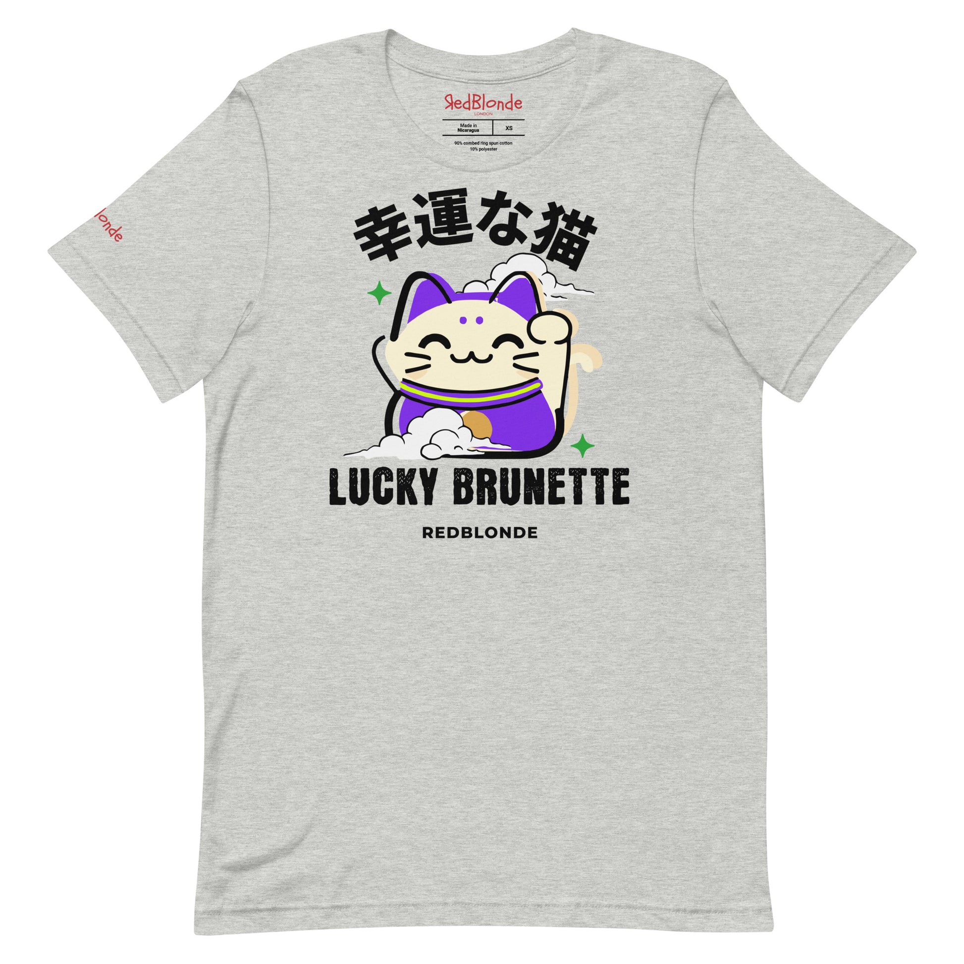 A light heather gray unisex T-shirt with a classic crew neckline and short sleeves. The front features a colorful graphic of a waving Maneki-neko (lucky cat) in cream and purple, surrounded by small green sparkles. Above the cat, bold black Japanese characters are displayed. Below the cat, the text reads “LUCKY BRUNETTE” in black block letters, with “REDBLONDE” printed underneath in smaller font. Branded RedBlonde in red inside neck and right sleeve