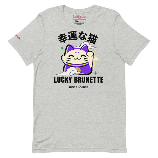 A light heather gray unisex T-shirt with a classic crew neckline and short sleeves. The front features a colorful graphic of a waving Maneki-neko (lucky cat) in cream and purple, surrounded by small green sparkles. Above the cat, bold black Japanese characters are displayed. Below the cat, the text reads “LUCKY BRUNETTE” in black block letters, with “REDBLONDE” printed underneath in smaller font. Branded RedBlonde in red inside neck and right sleeve