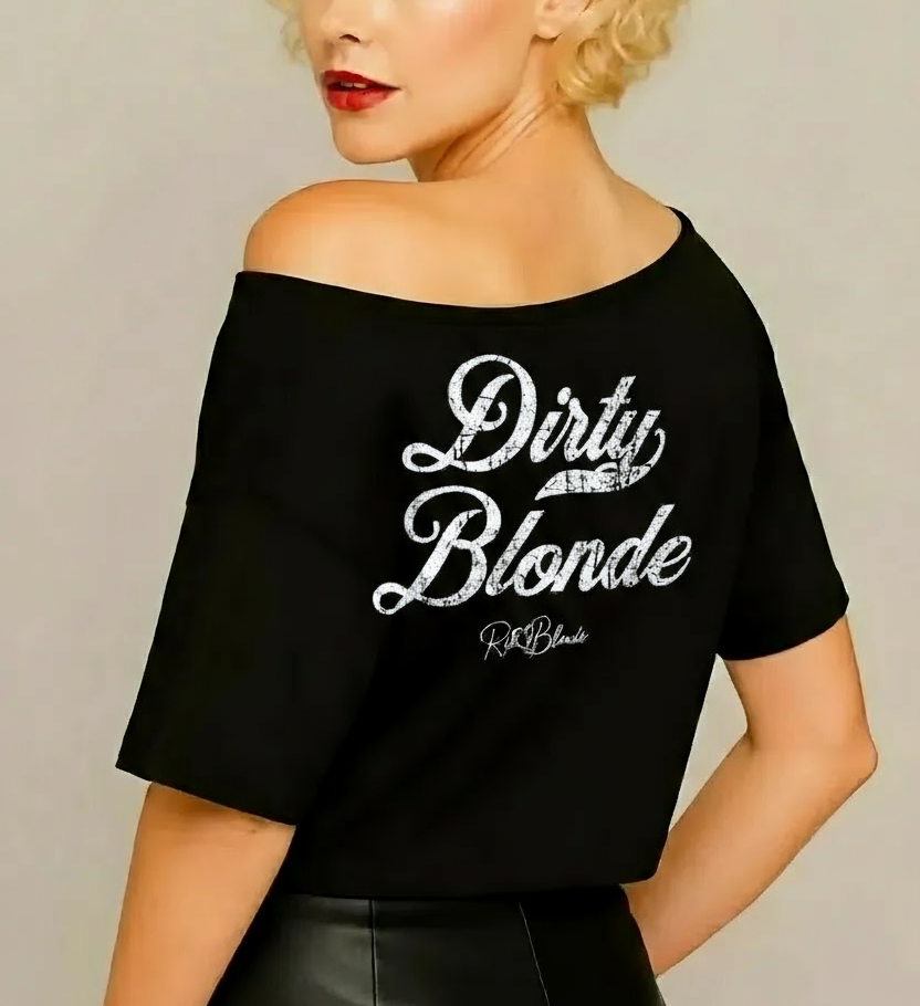 Blonde wears, seen from back view, a black asymmetric off-the-shoulder waist length t-shirt featuring the bold white distressed Milkstore font “Dirty Blonde” with the RedBlonde logo printed below. The relaxed, oversized fit and one-shoulder neckline give it a modern, effortless look.
