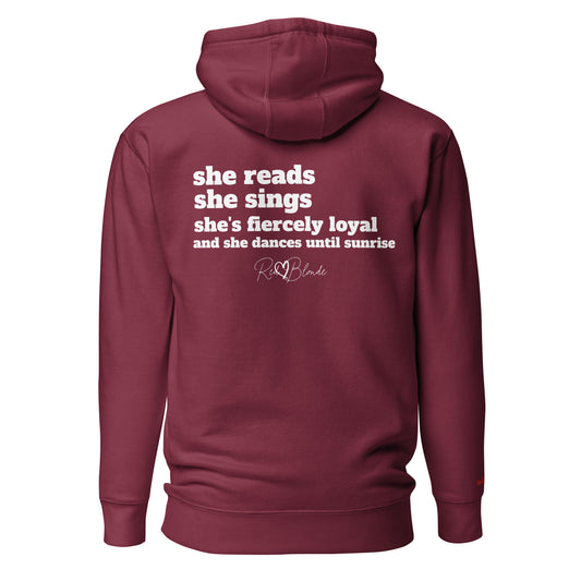 Back view of a maroon unisex hoodie featuring white text that reads, “she reads, she sings, she’s fiercely loyal, and she dances until sunrise,” with the RedBlonde logo printed underneath. The design conveys confidence and personality with a bold, uplifting message.