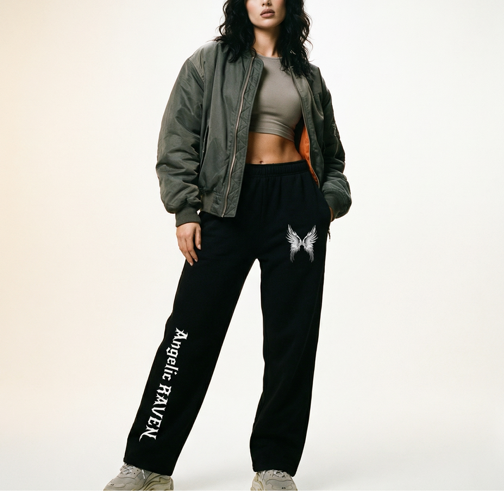Model wears black straight-leg joggers with white drawstrings, featuring a small white angel-wing graphic on the upper left thigh and ‘Angelic Raven’ printed vertically down the right leg in New Rocker gothic style lettering, shown flat against a white background.