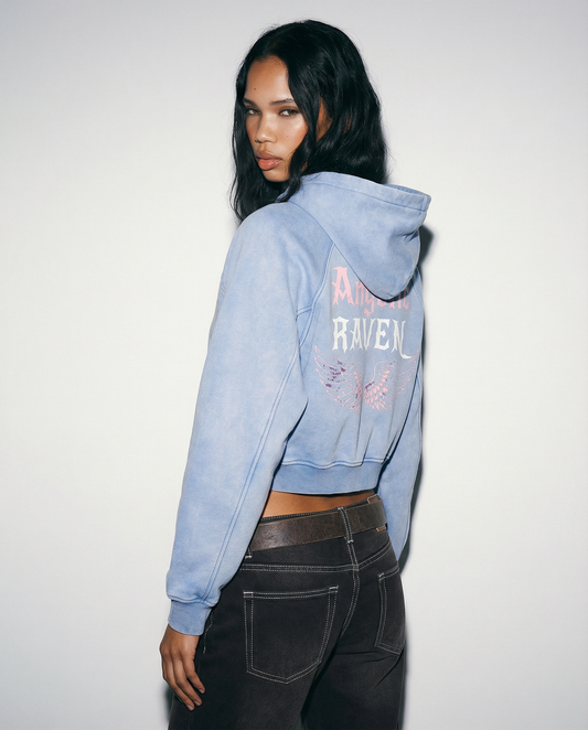 Model wears a pale blue cropped hoodie with hood up, featuring “Angelic Raven” New Rocker font text and iridescent angel wings graphic printed across the back on a white background.