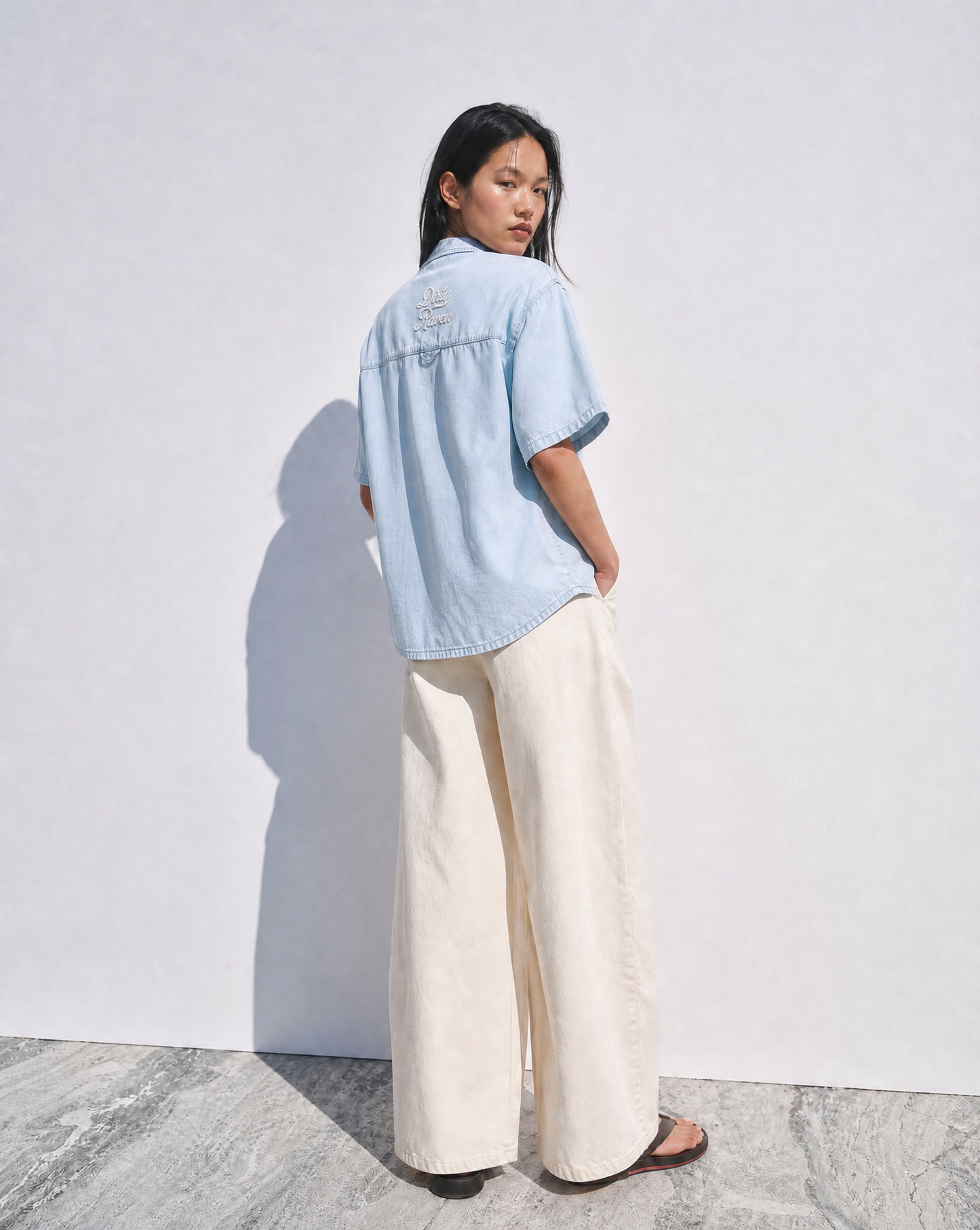 models wears a  light blue, short-sleeve button-up shirt with a subtle faded finish and a small “Dirty Raven” Milkstore script logo centered across the upper back.