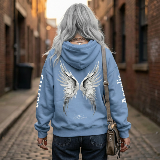 model wears a blue hoodie featuring large silver angel wings printed across the back, with black RedBlonde signature logo underneath. Each sleeve features gothic New Rockstar font graphic text  one sleeve says 'Angelic' and the other 'Silver' 