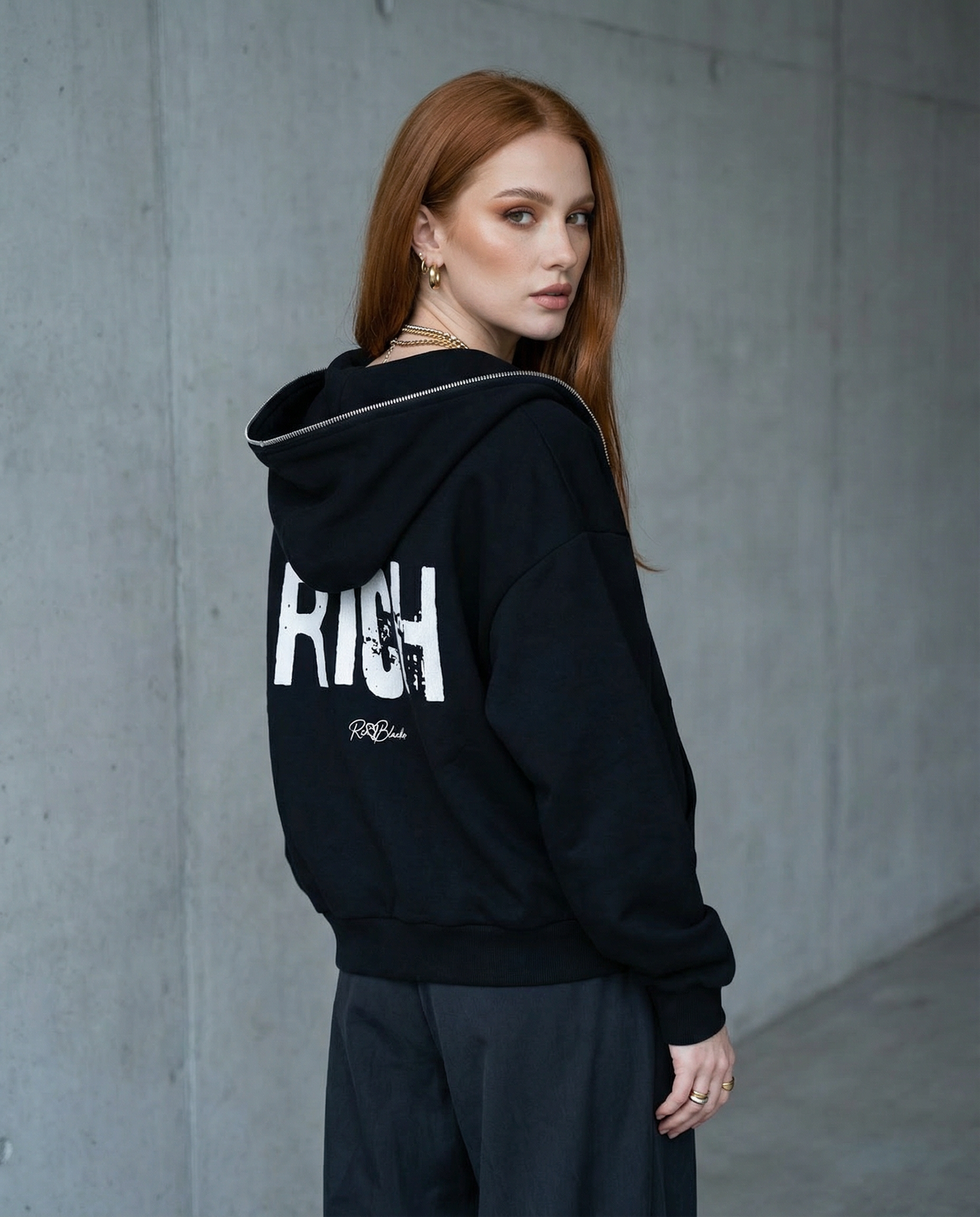 model wears black boxy zip-up hoodie with a large white distressed “RICH” in Ed Gein graphic across the back and a small RedBlonde logo underneath.