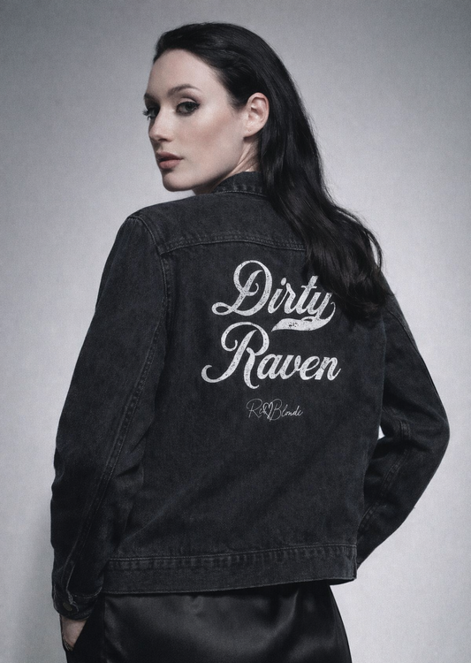 The back of a washed-black denim jacket showing large white embroidered Miklstore font script reading "Dirty Raven" with the RedBlonde signature below. The jacket has a relaxed, slightly cropped silhouette and long sleeves with button cuffs.