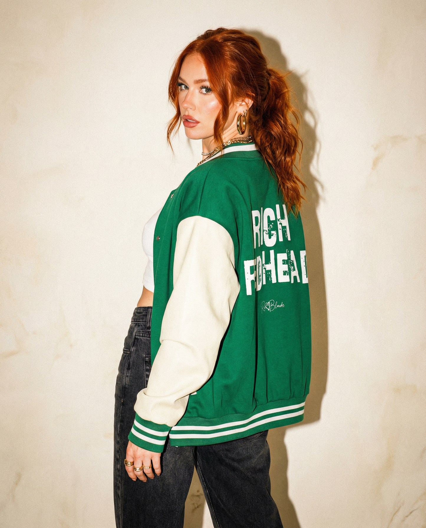 model wears a green & white varsity-style jacket with cream sleeves and striped ribbed trims. The back features large white text reading “RICH REDHEAD” in Ed Gein font with a small RedBlonde signature logo in the centre between the words