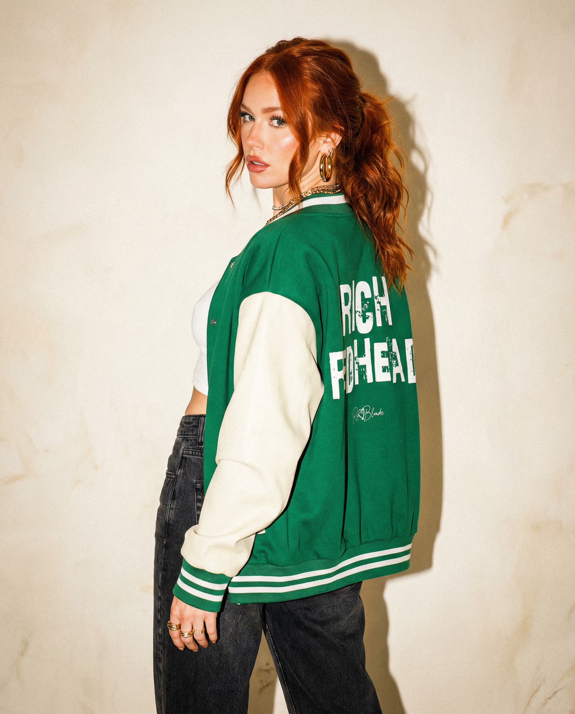 model wears a green & white varsity-style jacket with cream sleeves and striped ribbed trims. The back features large white text reading “RICH REDHEAD” in Ed Gein font with a small RedBlonde signature logo in the centre between the words