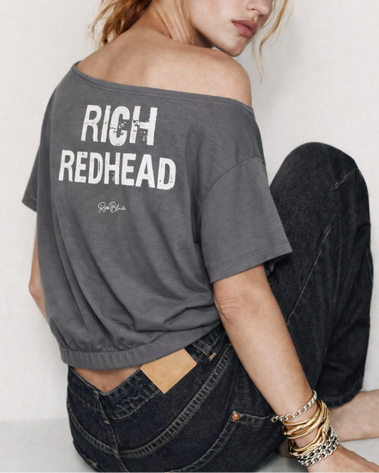 model wears a grey asymmetrical shoulder t-shirt with bold white Ed Gein font “RICH REDHEAD" slogan on the front and RedBlonde logo beneath.