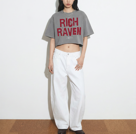 model wears a grey cropped T-shirt with oversized short sleeves and a red “RICH RAVEN” in Ed Gein font graphic on the front, plus a small RedBlonde script logo underneath.