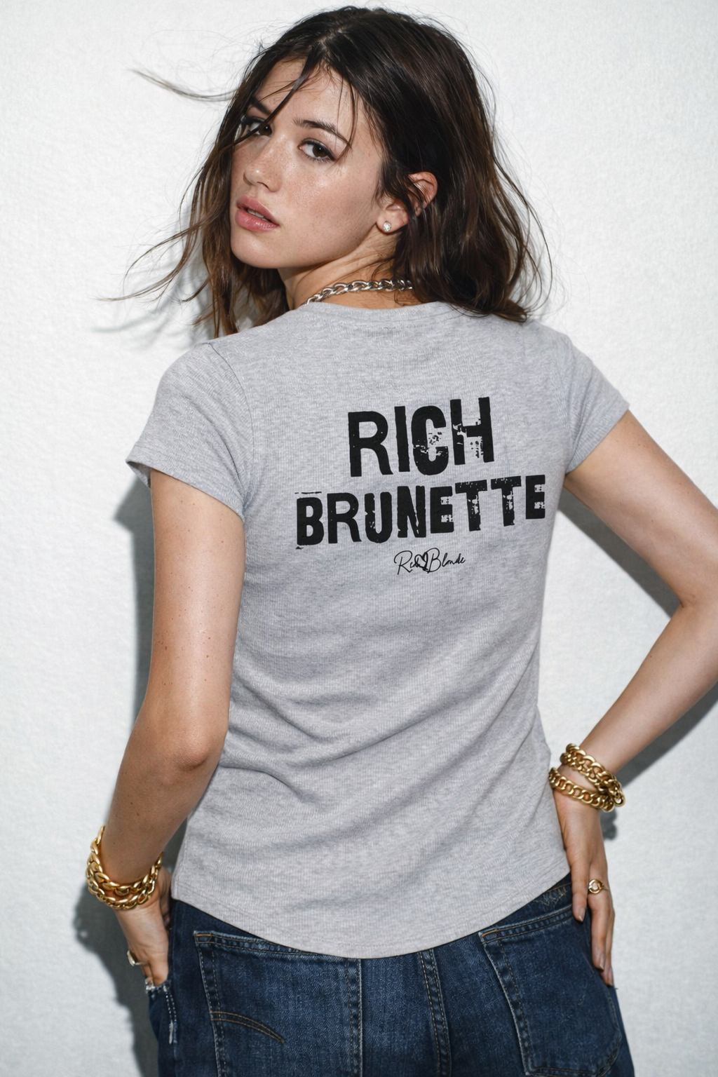 model wears a light grey slightly fitted women’s henley T-shirt with short sleeves and a gently curved hem. Centered on the upper back is bold distressed black Ed Gein text reading “RICH BRUNETTE” with a small RedBlonde signature script logo beneath it. The shirt is shown on a plain white background.