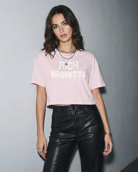 model wears a orchid crop top with a relaxed fit, short sleeves, and a wide crew neckline. The front features bold black distressed Ed Gein lettering reading “RICH BRUNETTE” with the RedBlonde signature logo underneath in black