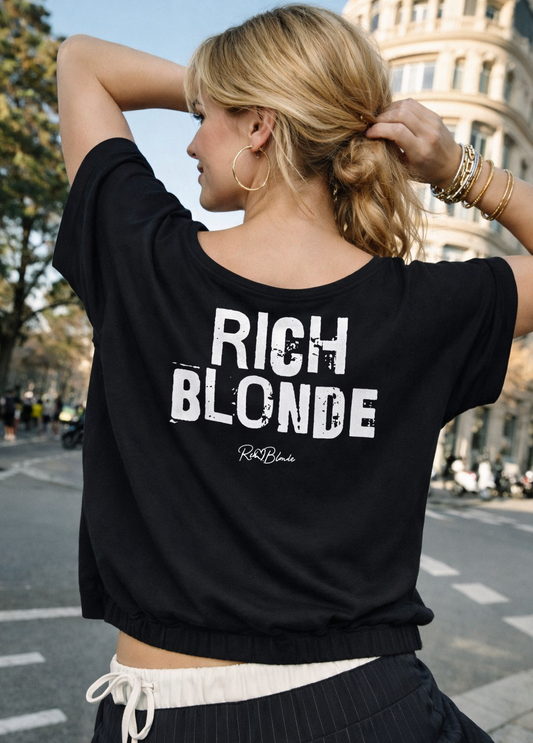model wears a black asymmetrical shoulder t-shirt with bold black Ed Gein font “RICH BLONDE" slogan on the front and RedBlonde logo beneath.
