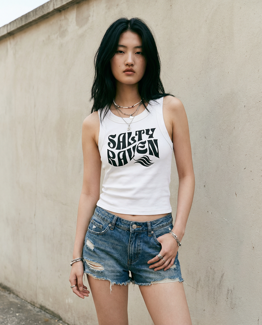 model wears a white sleeveless crop tank top with a high neckline, featuring a black Solino Display retro-style “Salty Raven” font graphic across the chest 