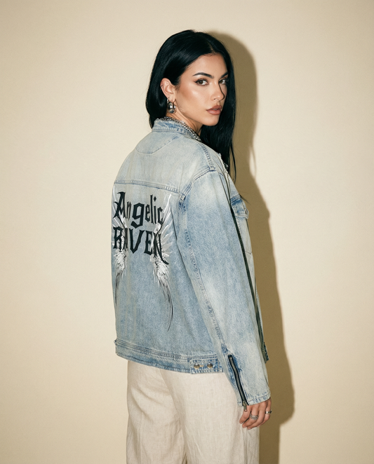 model wears a light-wash denim jacket shown from the back on a white background. The sleeves have zippered cuffs, and the back features a large graphic with white angel wings and black gothic New Rocker style text reading “Angelic RAVEN.”