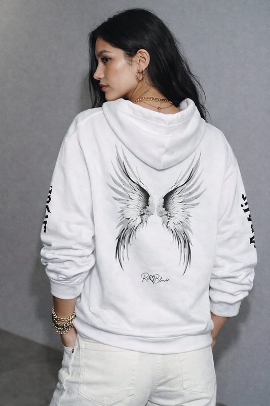 model wears a white oversize hoodie featuring large silver angel wings printed across the back, with white RedBlonde signature logo underneath. Each sleeve features gothic New Rockstar font graphic text  one sleeve says 'Angelic' and the other 'Raven' 