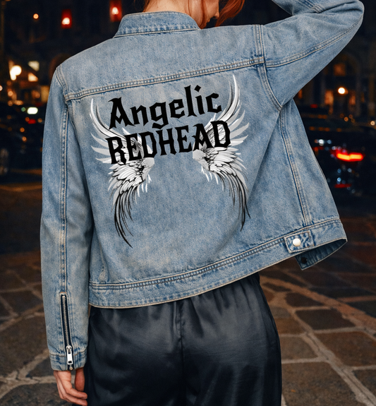 model wears a light-wash denim jacket shown from the back on a white background. The sleeves have zippered cuffs, and the back features a large graphic with white angel wings and black gothic New Rocker style text reading “Angelic REDHEAD.”
