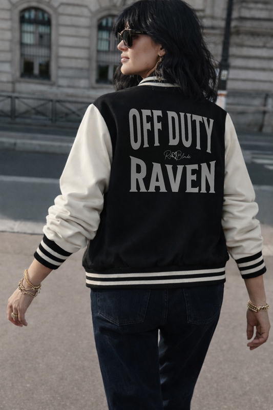 model wears a Black & white varsity-style jacket with cream sleeves and striped ribbed trims. The back features large white text reading “OFF DUTY RAVEN” in Brookfield font with a small RedBlonde signature logo in the centre between the words
