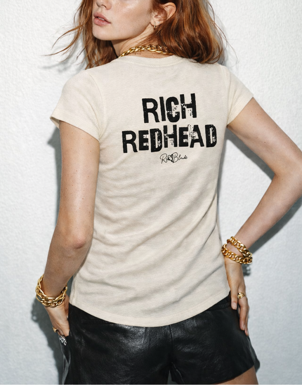 model wears a  beige slightly fitted women’s henley T-shirt with short sleeves and a gently curved hem. Centered on the upper back is bold distressed black Ed Gein text reading “RICH REDHEAD” with a small RedBlonde signature script logo beneath it. 