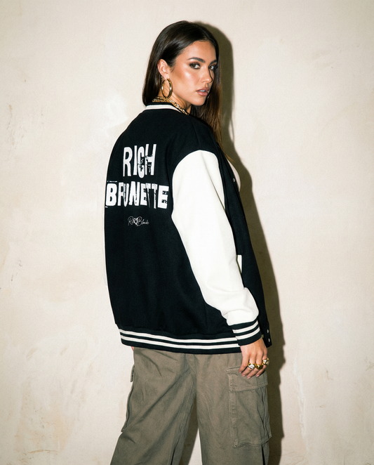 model wears a black & white varsity-style jacket with cream sleeves and striped ribbed trims. The back features large white text reading “RICH BRUNETTE” in Ed Gein font with a small RedBlonde signature logo in the centre between the words