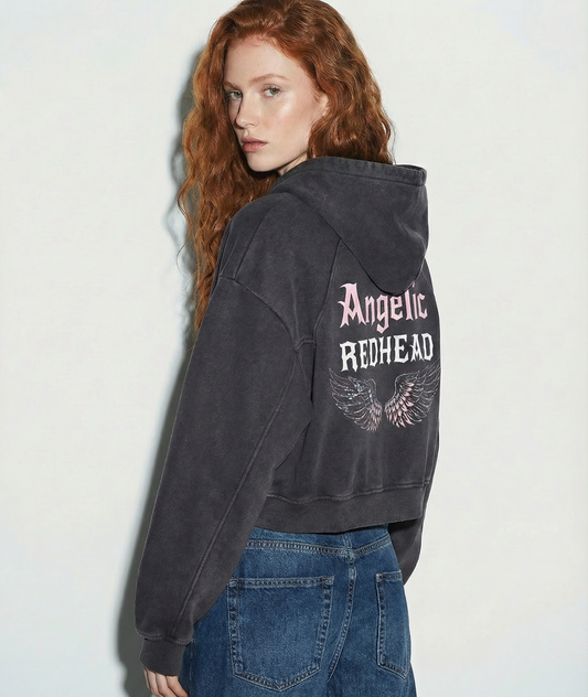 model wears black cropped hoodie with hood up, featuring “Angelic Redhead” New Rocker font text and iridescent angel wings graphic printed across the back on a white background.