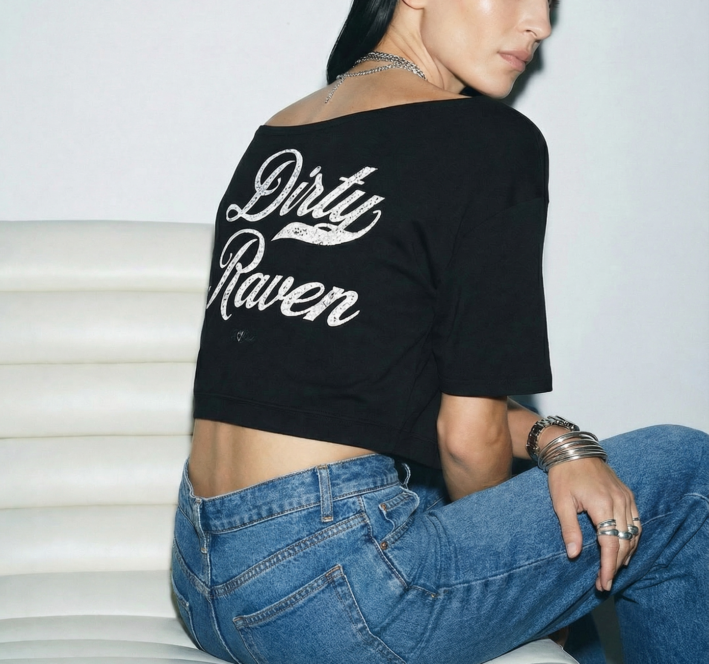 Back view - model wears a  Black asymmetric off-the-shoulder waist length t-shirt featuring the bold white distressed Milkstore font “Dirty Raven” with the RedBlonde logo printed below. The relaxed, oversized fit and one-shoulder neckline give it a modern, effortless look.