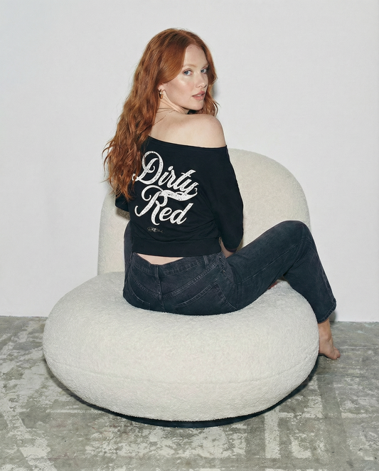 Back view of a model wearing a black asymmetric off-the-shoulder waist length t-shirt featuring the bold white distressed Milkstore font “Dirty Red” with the RedBlonde logo printed below. The relaxed, oversized fit and one-shoulder neckline give it a modern, effortless look.
