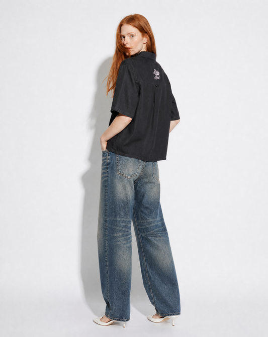 model wears a black, short-sleeve button-up shirt with a subtle faded finish and a small “Dirty Red” Milkstore script logo centered across the upper back.