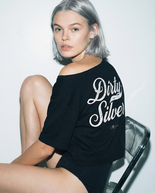  Back view - model wears black asymmetric off-the-shoulder waist length t-shirt featuring the bold white distressed Milkstore font “Dirty Silver” with the RedBlonde logo printed below. The relaxed, oversized fit and one-shoulder neckline give it a modern, effortless look.