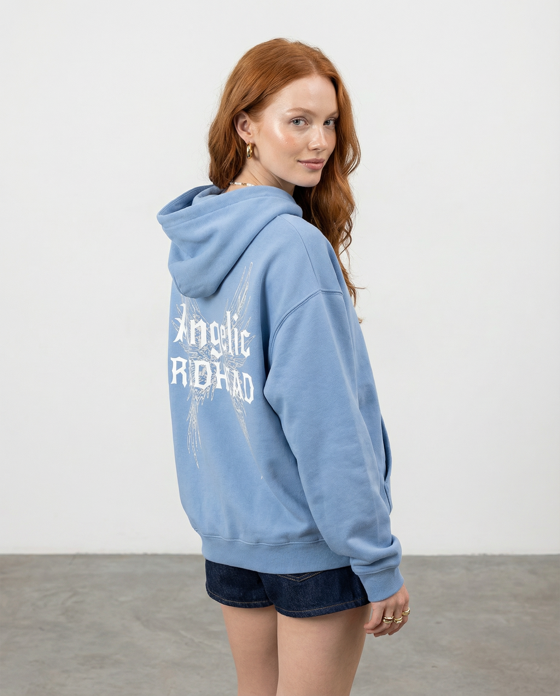 model wears light blue oversized fleeced hoodie with a back print featuring white gothic “Angelic Redhead” text layered over large angel wing artwork.