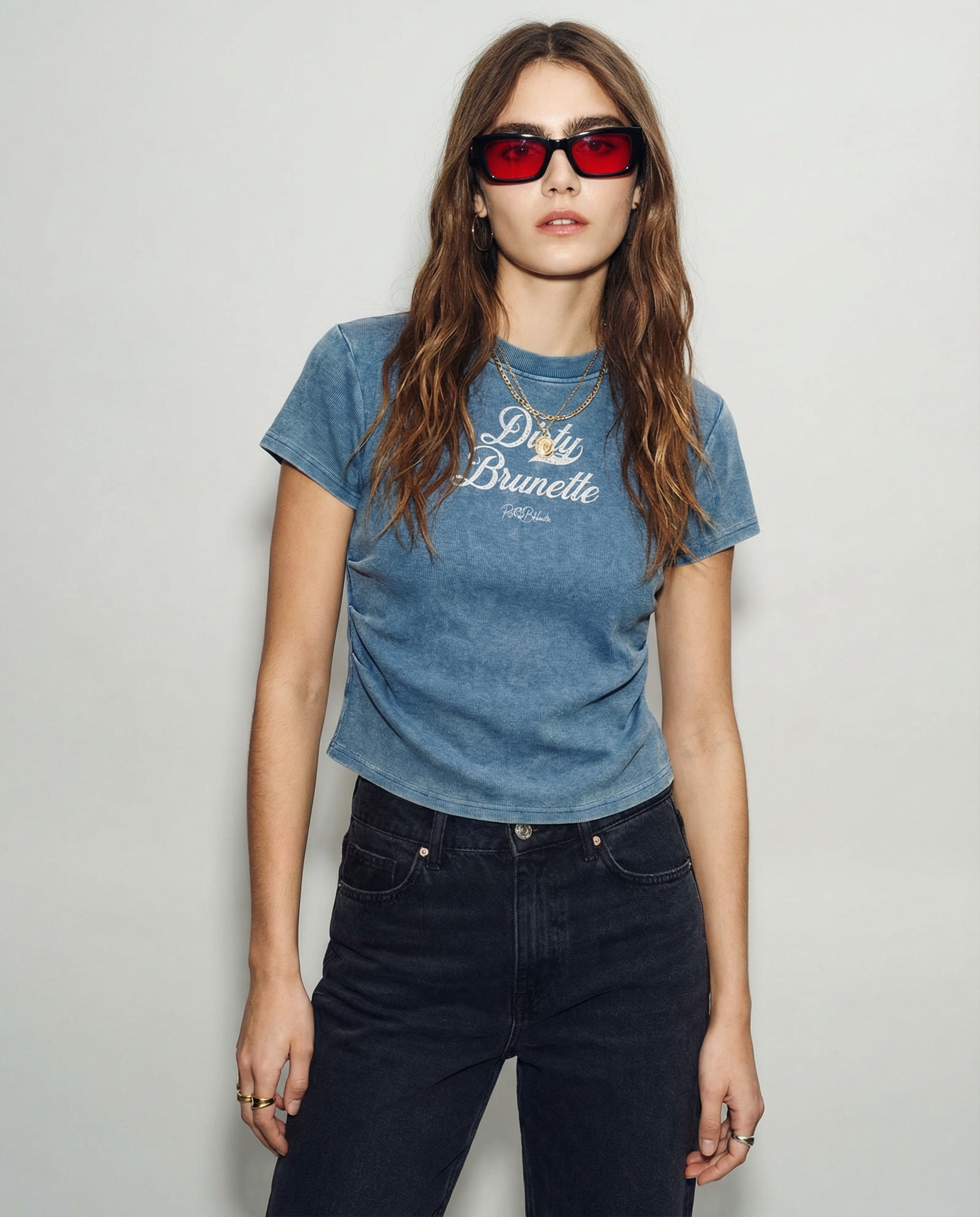 A model wears a fitted, short-sleeve blue crewneck T-shirt laid flat on a white background. It has subtle ruching/contouring at the sides and a slightly curved hem. White script text across the chest reads “Dirty Brunette,” in Milkstore 01 script with a small cursive “RedBlonde” logo beneath.