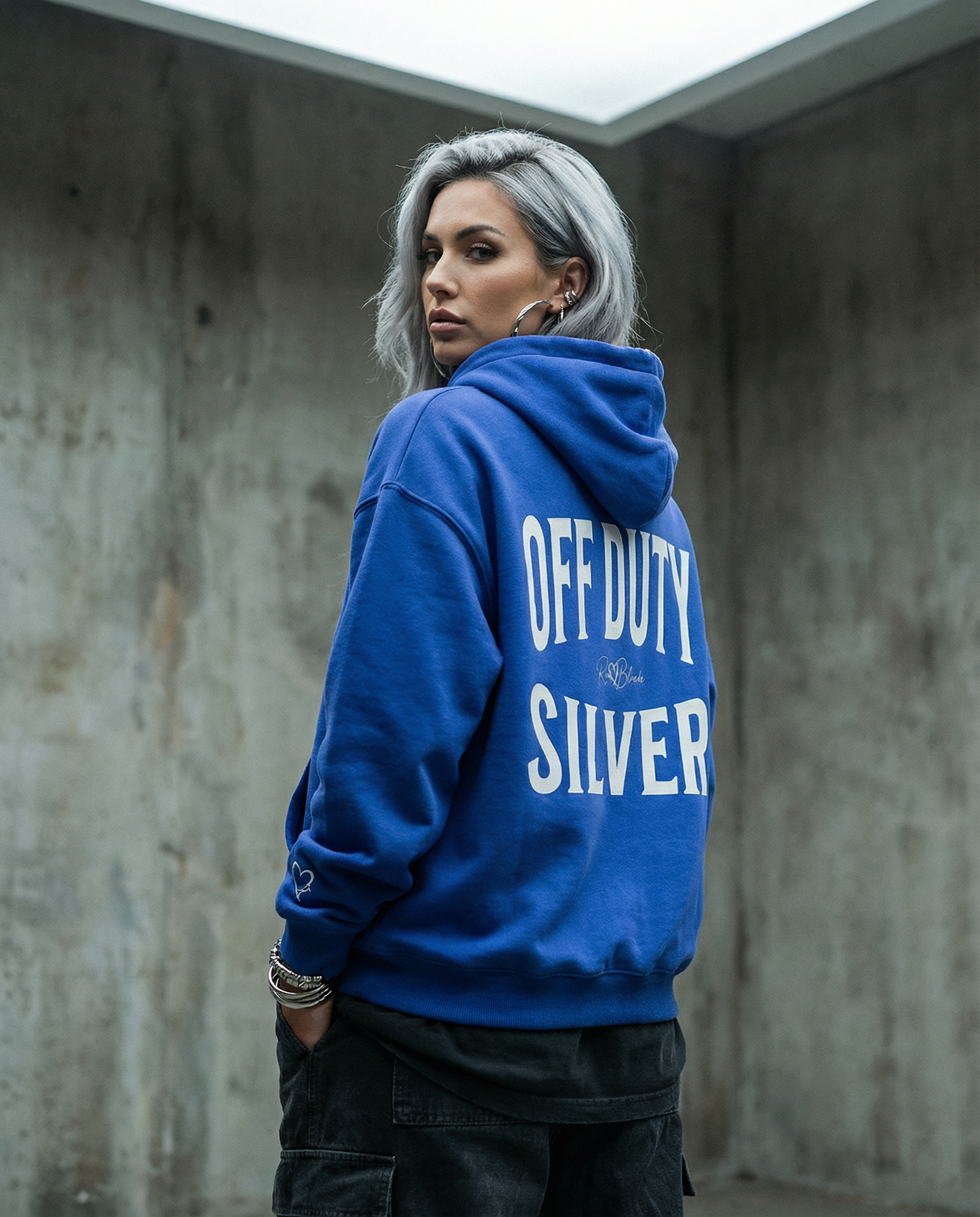 Model wears a blue hoodie, back view, with large white  ‘OFF DUTY SILVER' Brookfield text print and small RedBlonde signature logo beneath. white heart detail on the sleeve