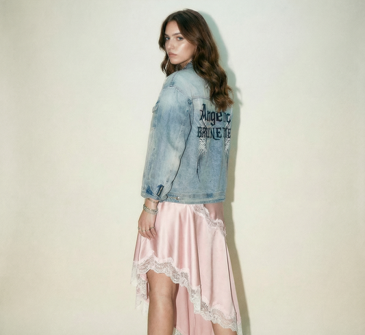 model wears a light-wash denim jacket shown from the back on a white background. The sleeves have zippered cuffs, and the back features a large graphic with white angel wings and black gothic New Rocker style text reading “Angelic BRUNETTE.” styled with a pale pink satin skirt and white kitten sling back shoes 
