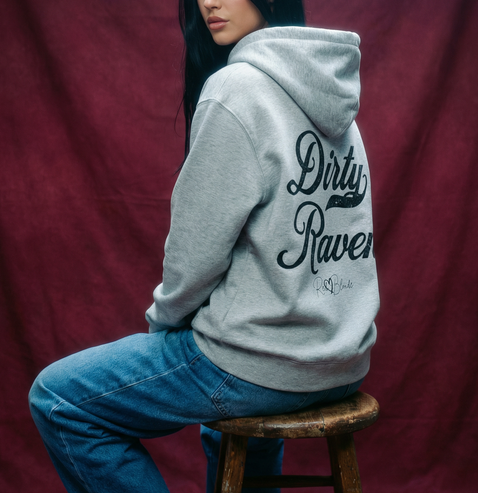 Raven haired Model wears a grey coloured hoodie with bold white Milkstore vintage-style script text “Dirty Raven” across the back and a subtle RedBlonde signature logo underneath.