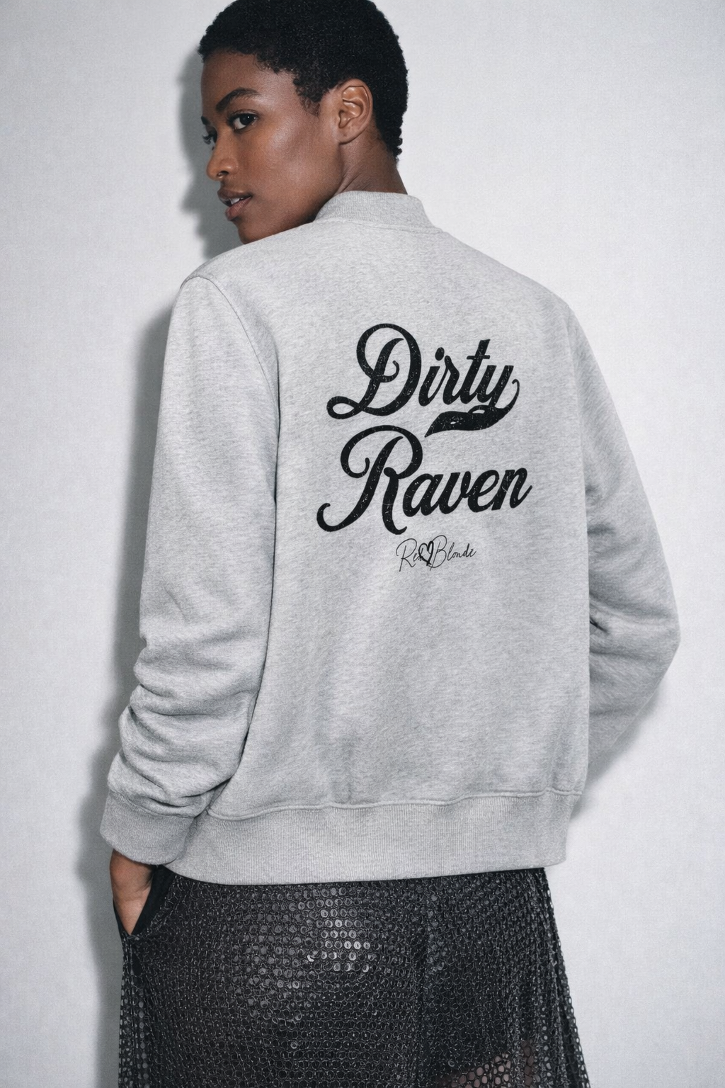 black haired model wears light grey crewneck zip up varsity jacket  laid flat on a white background. It has ribbed cuffs, collar, and hem. Large black Milkstore 01 script text across the back reads “Dirty Raven,” with a small cursive RedBlonde logo beneath.