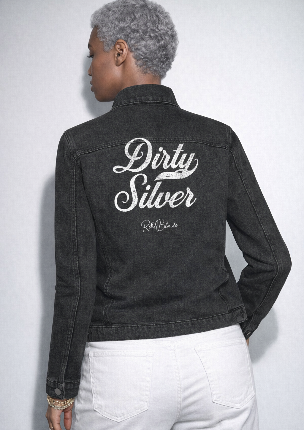 model wears washed-black denim jacket showing large white embroidered Milkstore font script reading "Dirty Silver" with the RedBlonde signature below. The jacket has a relaxed, slightly cropped silhouette and long sleeves with button cuffs.
