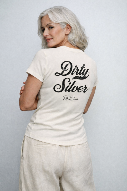 model wears a short-sleeve off white T-shirt laid flat on a white background. It features large black Milkstore 01 script text reading “Dirty Silver” across the upper back, with a small cursive “RedBlonde” logo beneath. The hem is slightly curved.
