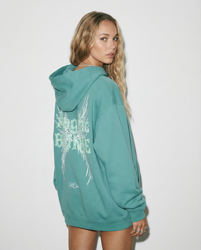 model wears a teal hoodie with a large distressed winged “Angel Blonde” graphic in white and mint across the upper back, plus a small RedBlonde script logo near the lower back hem.