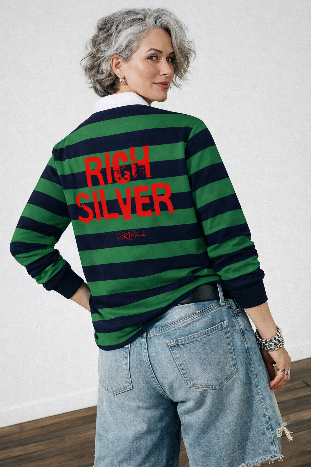 model wears a long-sleeved rugby polo shirt featuring a pattern of alternating horizontal green and navy stripes. The center of the back displays the words "RICH SILVER" in large, distressed Ed Gein red block lettering, with a smaller signature-style logo printed directly underneath. The shirt is finished with a solid white collar and matching white ribbed cuffs.