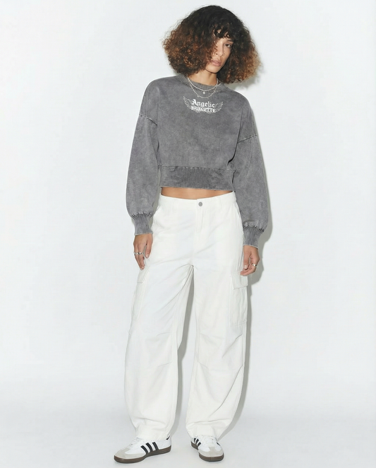 model wears grey mineral-wash cropped sweater with balloon sleeves, ribbed collar/cuffs/waistband, and a white “Angelic Brunette” wing graphic on the chest