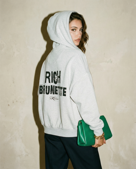 model wears a grey hem to hood zip-up hoodie with cropped oversized fit and ribbed cuffs/hem, featuring large distressed black “RICH BRUNETTE” Ed Gein text across the back with a small “RedBlonde” script logo beneath.