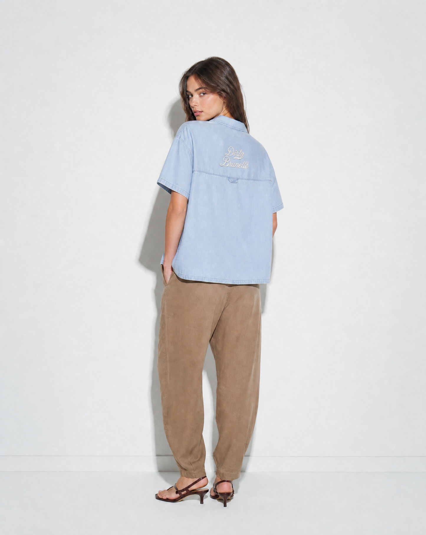 model wears light blue, short-sleeve button-up shirt with a subtle faded finish and a small “Dirty Brunette” Milkstore script logo centered across the upper back.