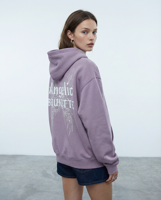 model wears lilac oversized fleeced hoodie with a back print featuring white gothic “Angelic Brunette” text layered over large angel wing artwork.