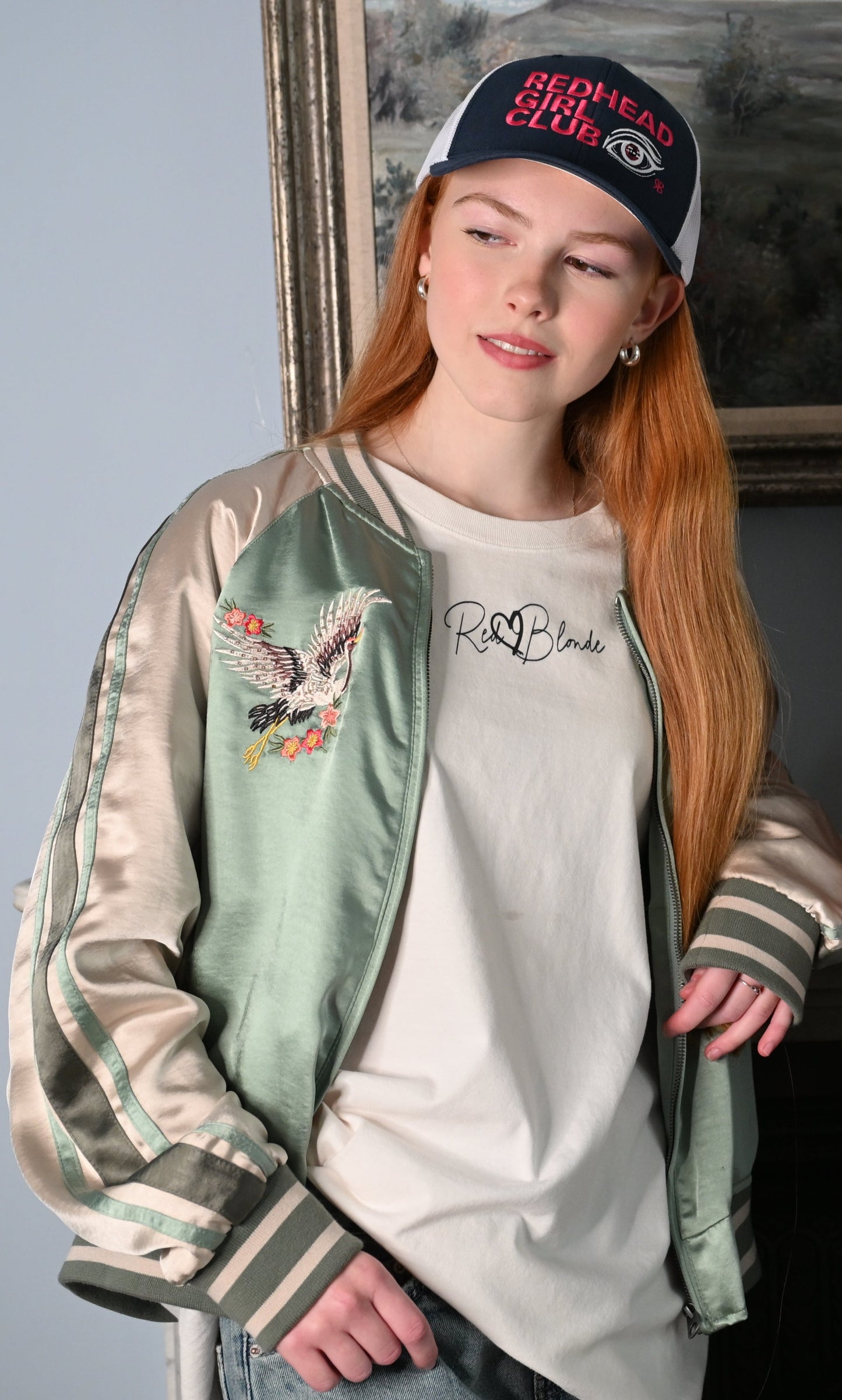 Redhead model wears navy/white mesh trucker hat featuring bold red embroidered text “Redhead Girl Club” alongside a detailed embroidered eye graphic and a small red RB logo.