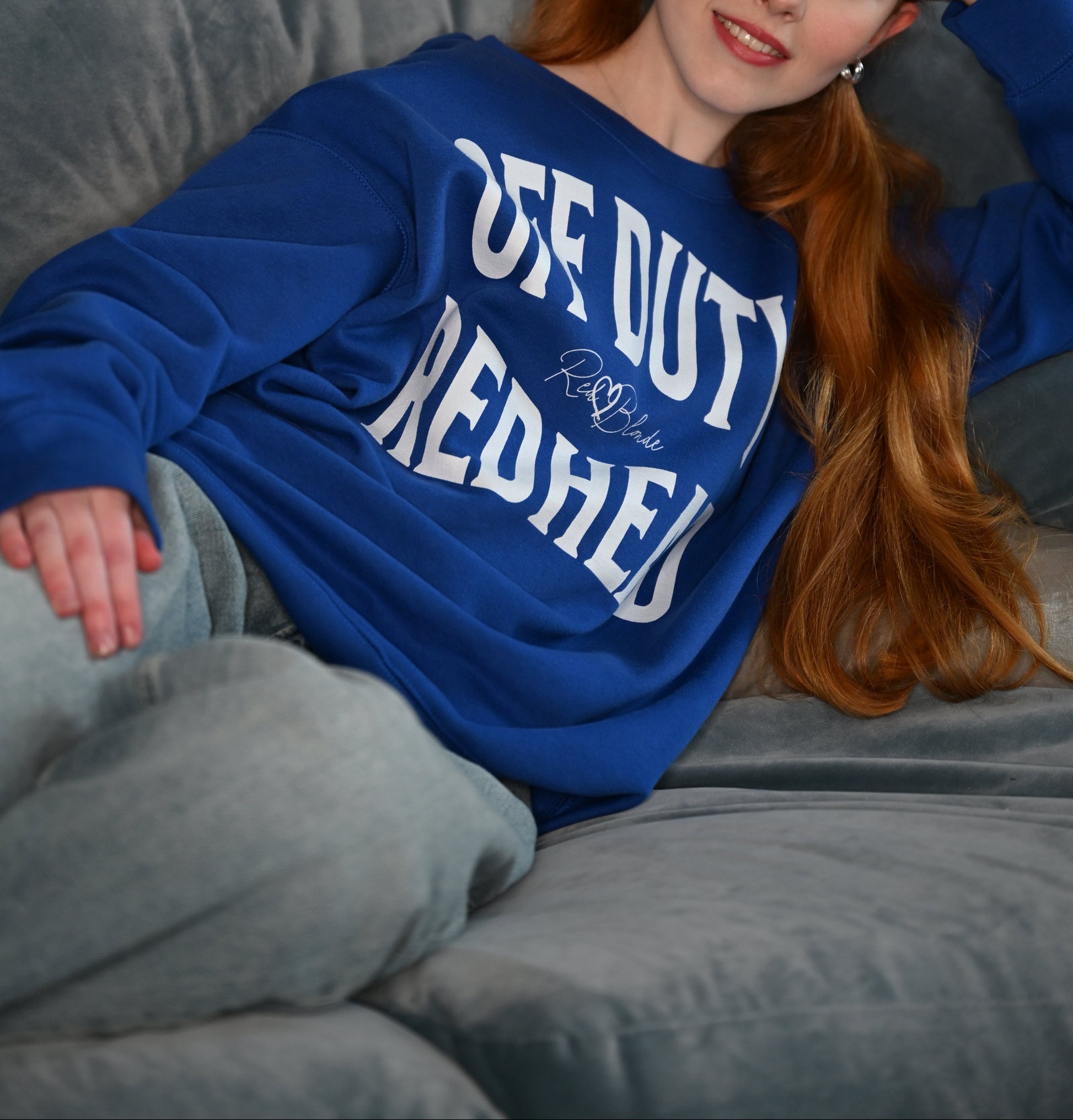A redhead wears a classic navy blue relaxed fit unisex sweatshirt with the words 'OFF DUTY REDHEAD' written in large Brookfield red font, across the front. Branded RedBlonde London in red on inside neck and right sleeve 