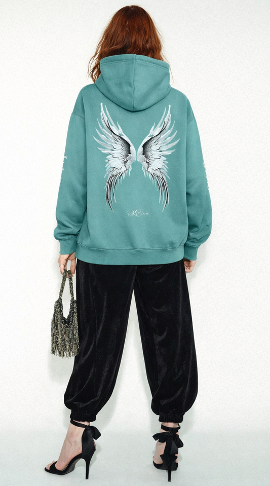 model wears a medium green hoodie featuring large silver angel wings printed across the back, with black RedBlonde signature logo underneath. Each sleeve features gothic New Rockstar font graphic text  one sleeve says 'Angelic' and the other 'Redhead' 