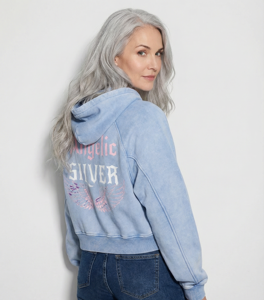 Back view - model wears a pale blue cropped hoodie with hood up, featuring “Angelic Silver” New Rocker font text and iridescent angel wings graphic printed across the back