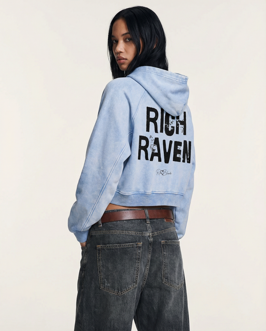 model wears  a cropped snow-wash pale blue hoodie with bold black “RICH RAVEN” Ed Gein font and the RedBlonde signature logo printed below.