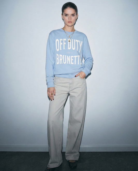 model wears pale blue crewneck sweatshirt with a large white “OFF DUTY BRUNETTE” graphic across the chest and a small RedBlonde script logo centered between the words.